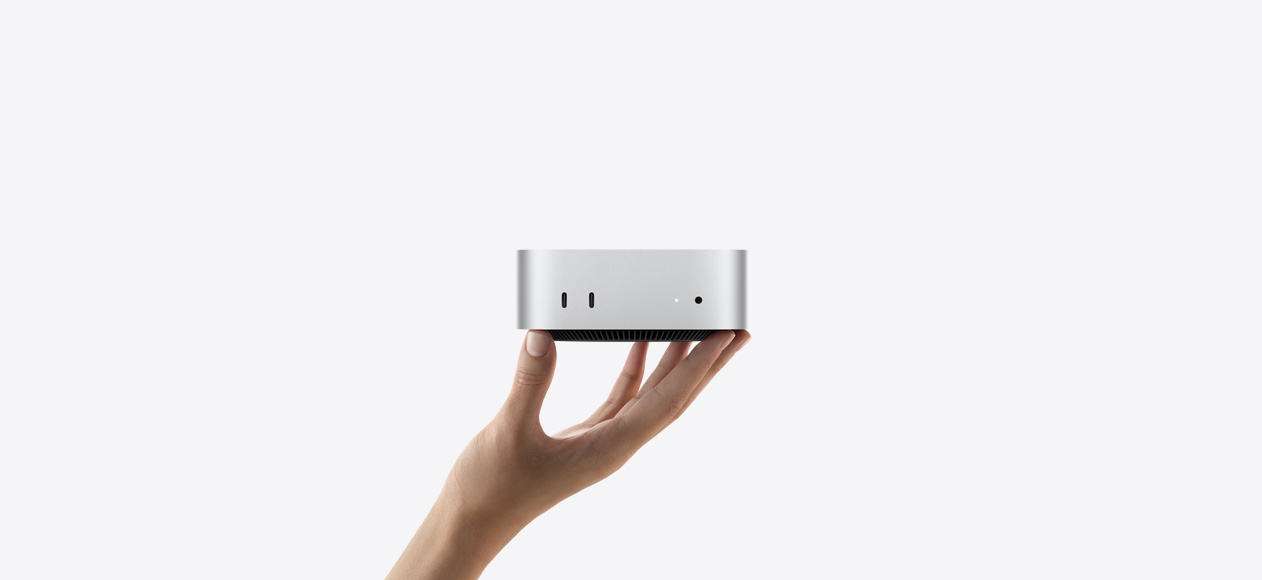 Widok z przodu na Maca mini leżącego na palcach wyciągniętej dłoni, na przodzie urządzenia widać dwa porty Thunderbolt, diodę statusu i gniazdo słuchawkowe, widać też czarną, zwężaną podstawę, płaski wierzch, zaokrąglone rogi i proste krawędzie, komputer w kolorze srebrnym